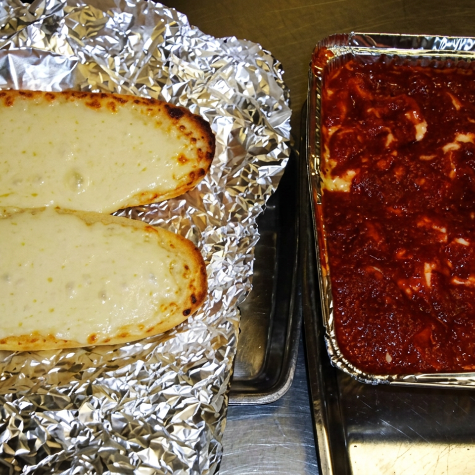 Two cheesy garlic bread loaves wrapped in foil beside a foil tray of red-sauce lasagna.