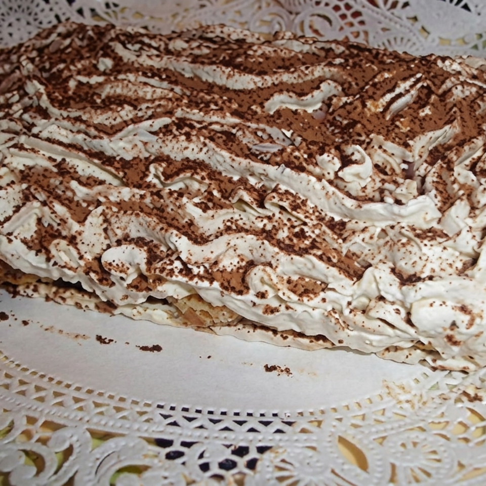 Rectangular tiramisu cake topped with whipped cream and cocoa powder, displayed on an ornate white doily-lined platter.
